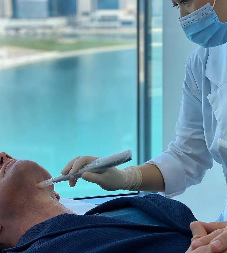 طبيب يرتدي قناعًا يطبّق جهازًا على شكل قلم على خدّ مريضة
